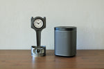 Load image into Gallery viewer, A piston clock made out of a Jaguar car&#39;s piston finished in gunmetal gray next to a speaker.
